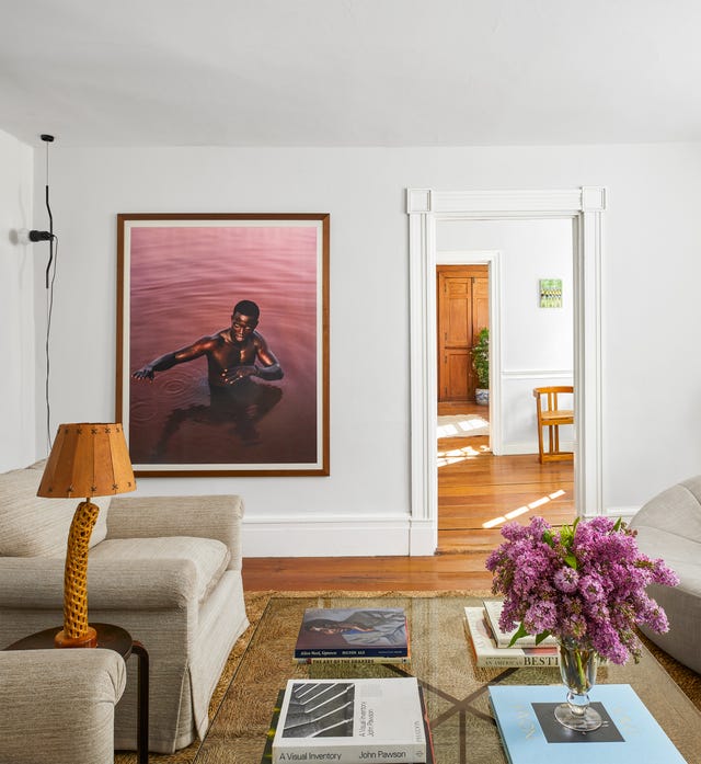 a sitting room has two upholstered chairs and a sofa in a similar beige fabric and a glass cocktail table with books and a vase of lilacs, a jute rug and a large framed photo of a man standing in water