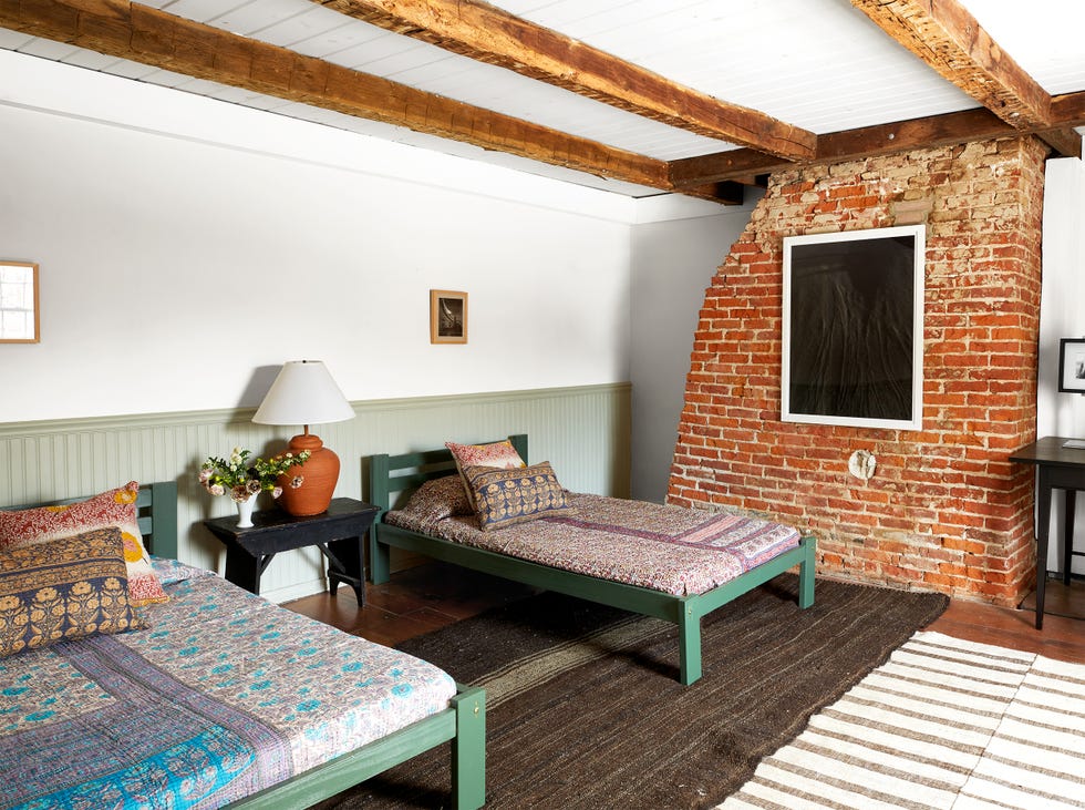 two twin beds in painted green wood frames with patterned coverlets and pillows, a table with a lamp between them, light green wainscoting on the wall behind and a brick wall with a large photo