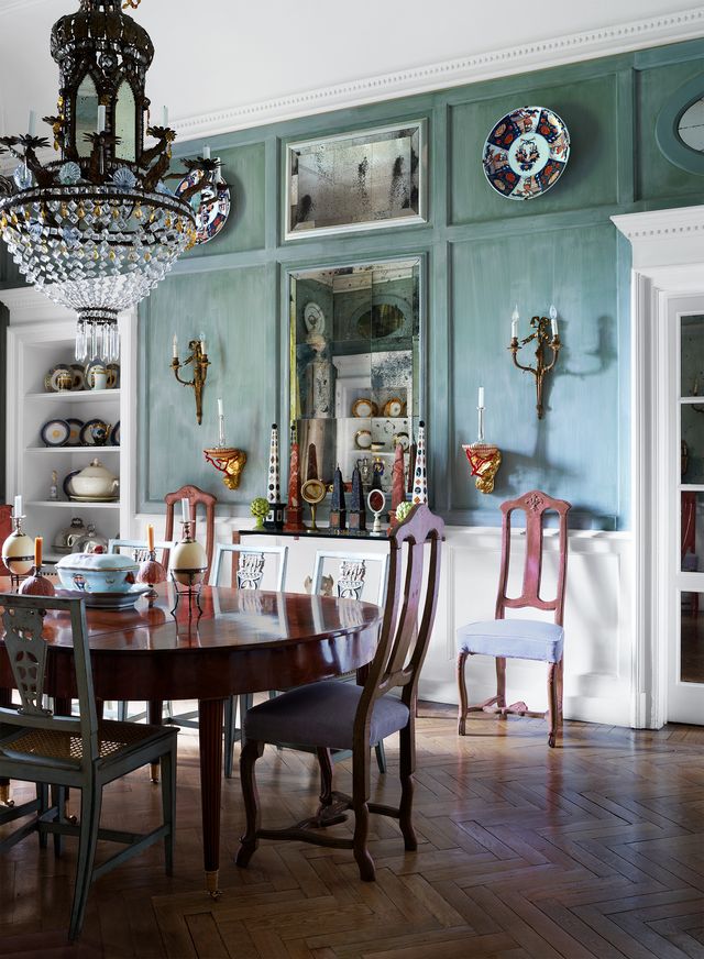 the dining room has a polished wood oblong table with numerous chairs and a glass chandelier above, pale green walls with antique sconces and candleholders, wall insets hold curios and vessels
