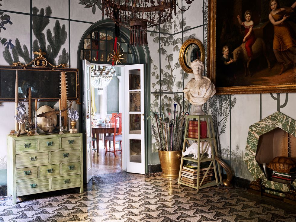 the entryway has a patterned marble floor, an antique chest of drawers with art objects on top, a stand with a marble bust of africa, a heptagonal fireplace, and an antique tin chandelier