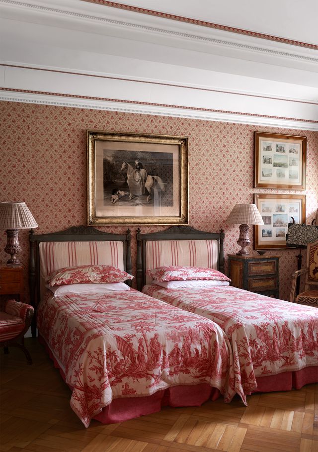 twin beds with wood striped fabric headboards and red and white toile de jouy patterned bedcovers, a mahogany side table and lamps beside each bed, the wall has framed photos and is sheathed in fabric