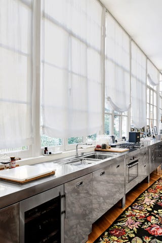 stainless steel kitchen cabinetry with multiple windows with white shades behind it and a dark flora rug on the floor