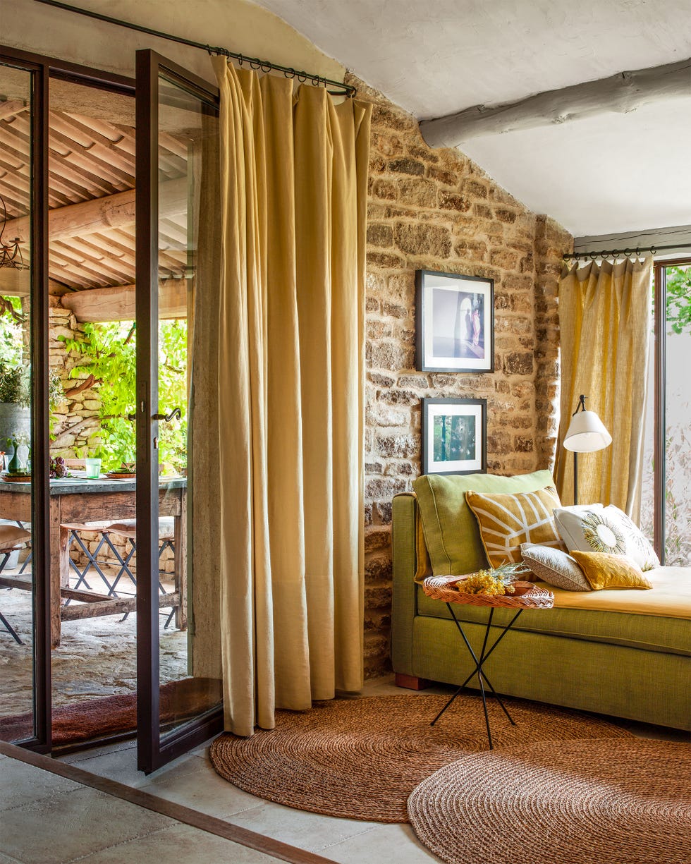 a sunroom has two round straw rugs, a sage green sofa with linen pillows and an accent table, two framed photos on a brick wall, gold linen curtains, and a large glass door leading to the outdoor dining area