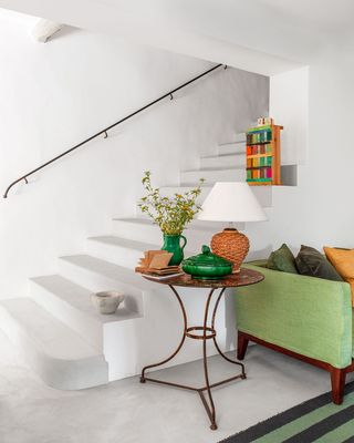a stairway has concrete stairs with a thin metal railing, an iron accent table with a straw lamp, an emerald green bowl and vase with flowers, and a sofa in a green fabric with pillows in fabrics of different colors