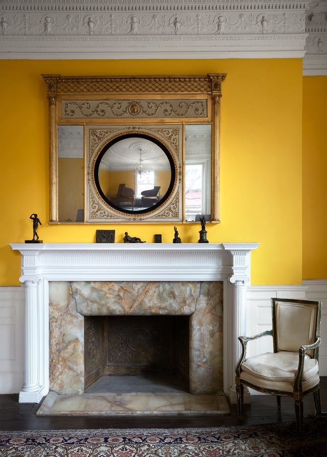 in a living room with yellow walls is an onyx fireplace with a painted white columned mantel with small sculptures atop and an armchair next to it, an ornate antique mirror above fireplace reflects two grand pianos