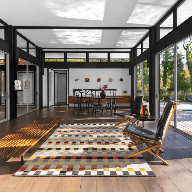 long  spare living room open to outside with sliding doors with a checkerboard rug at center and slatted wood furniture along it and a dining table and chairs at the far end