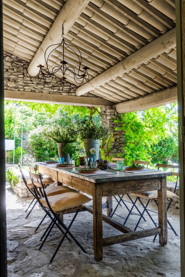 an outdoor dining area with a stone floor has a rustic rectangular table set for dining and six garden style chairs, above is a wrought iron chandelier and a ceiling make of cylindrical tiles