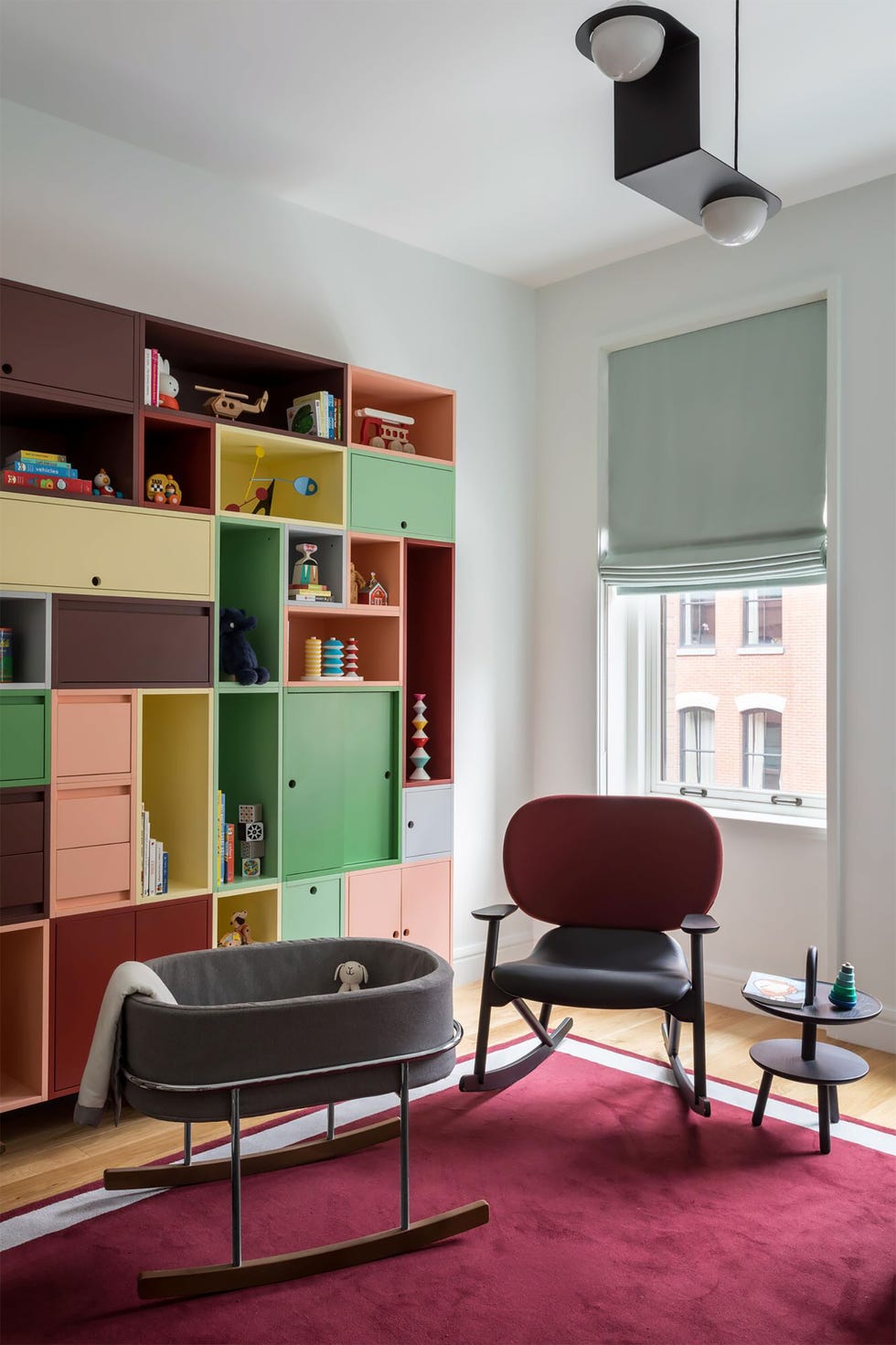 room with colorful cubbies a red rug and sleigh bassinet and matching chair and a light green roman shade on the window