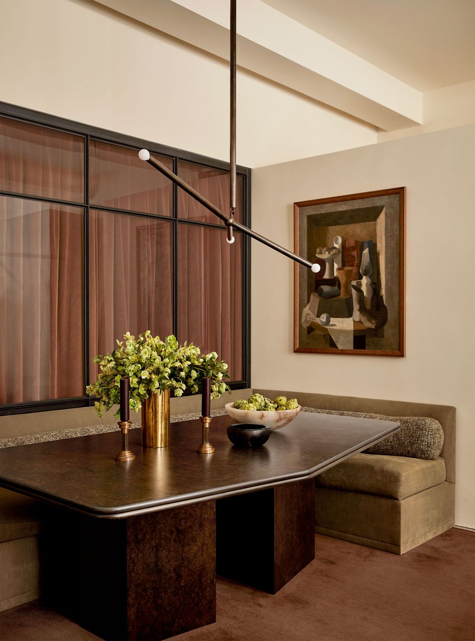 in a dining area is a five sided dark burl wood table against a frosted glass wall, a light brown cushioned banquette with an abstract artwork above one end, a three armed pendant, and a vase of green flowers