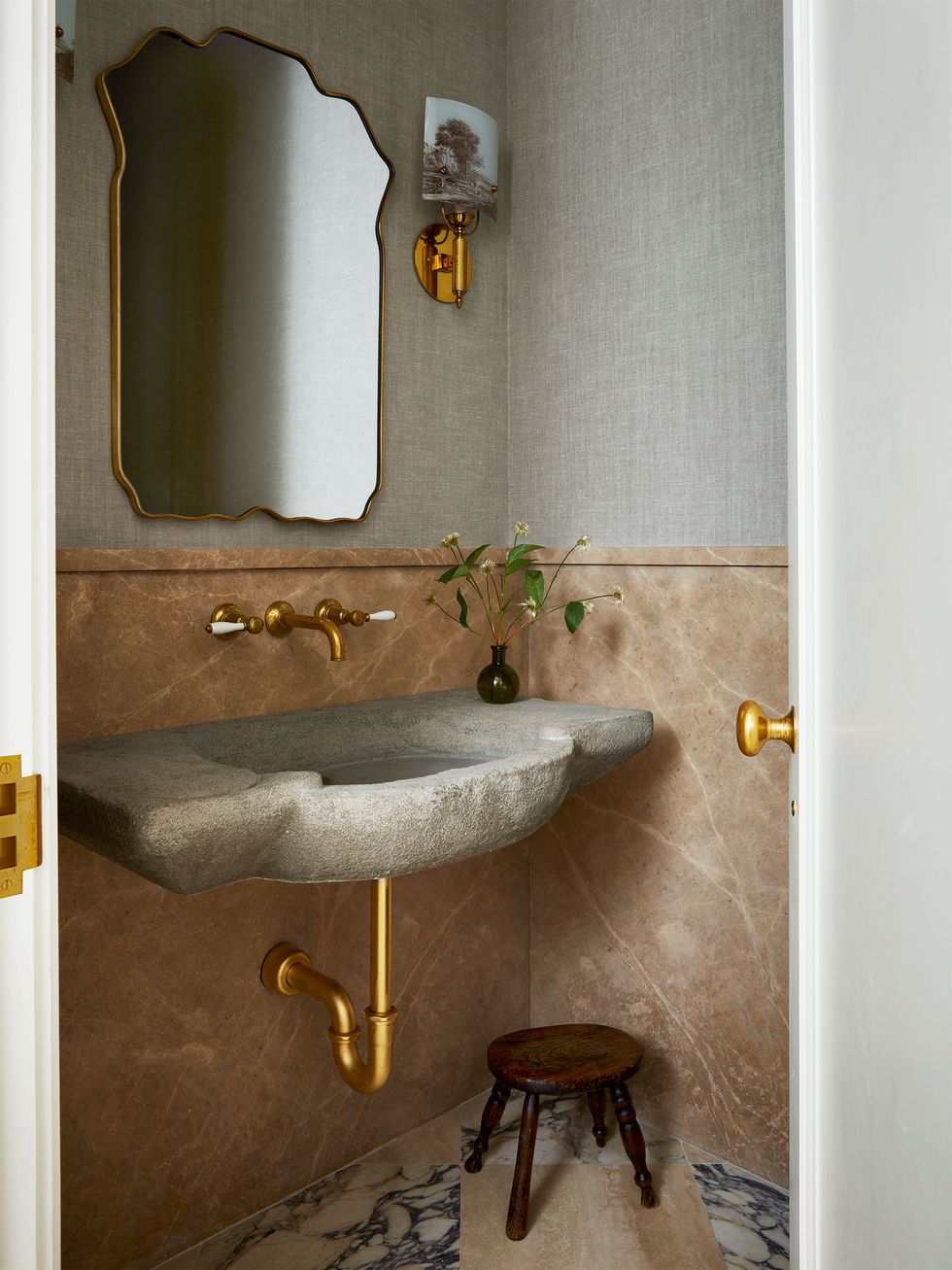a powder room has a marble floor and travertine on the bottom half of walls with wallpaper on upper half, a small wooden stool, a stone sink with copper fittings, antique tile sconces, and a wavy edged mirror