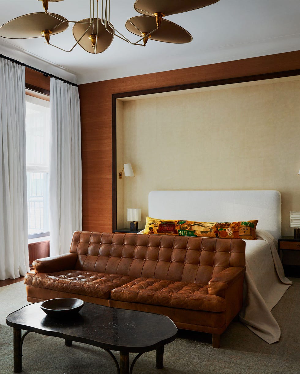 Augusta Hoffman - ELLE DECOR a primary bedroom has a wall inset with a bed with upholstered headboard, wall sconces and bedside lamps, a leather sofa at the foot, a table and a modern chair, multiarmed pendant, windows with drapes