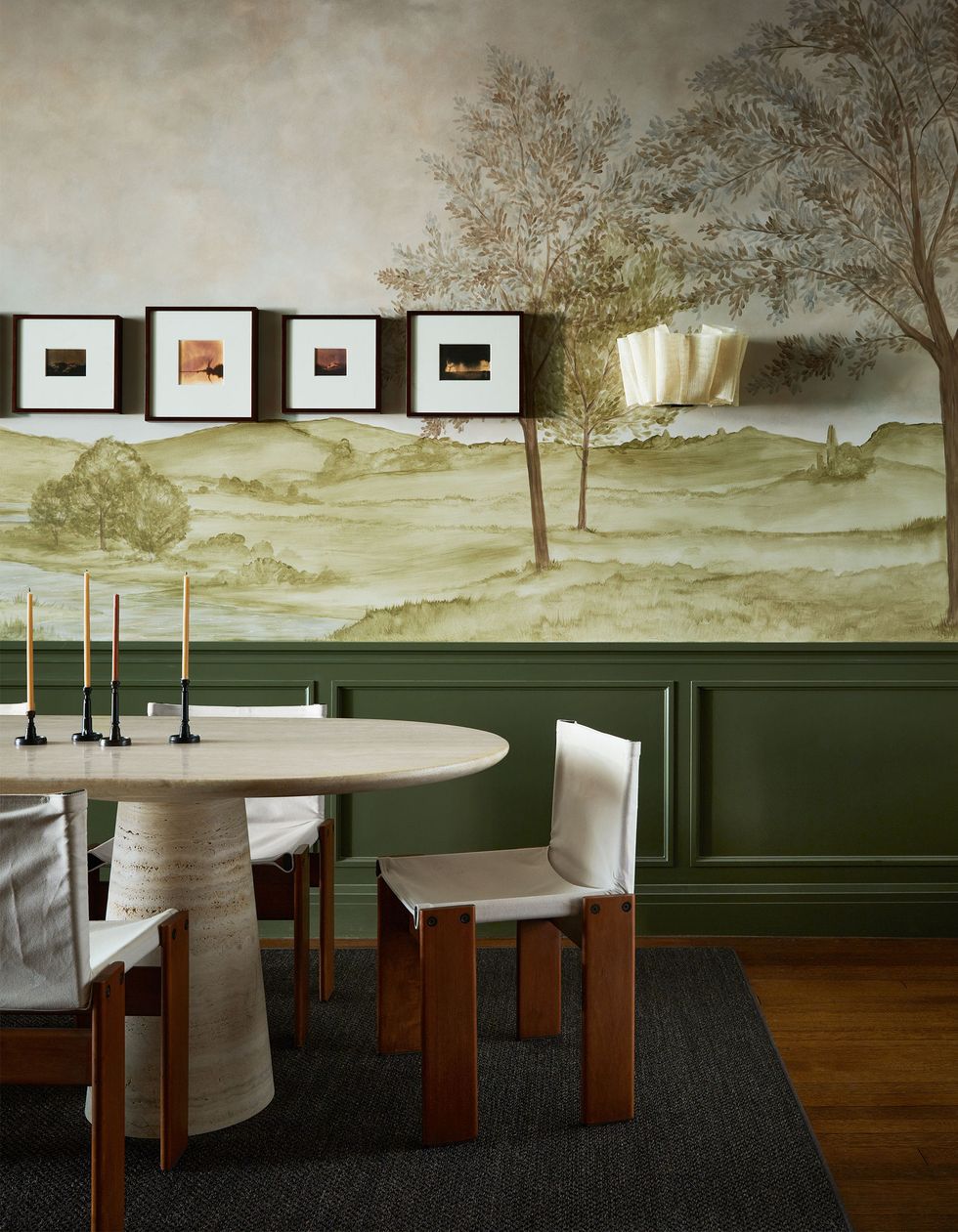the bottom of a dining room wall is dark olive and the top is a green toned mural of a rural scene, a ruffled wall sconce, small framed artworks, travertine table with pedestal legs, wood chairs with off white fabric seats