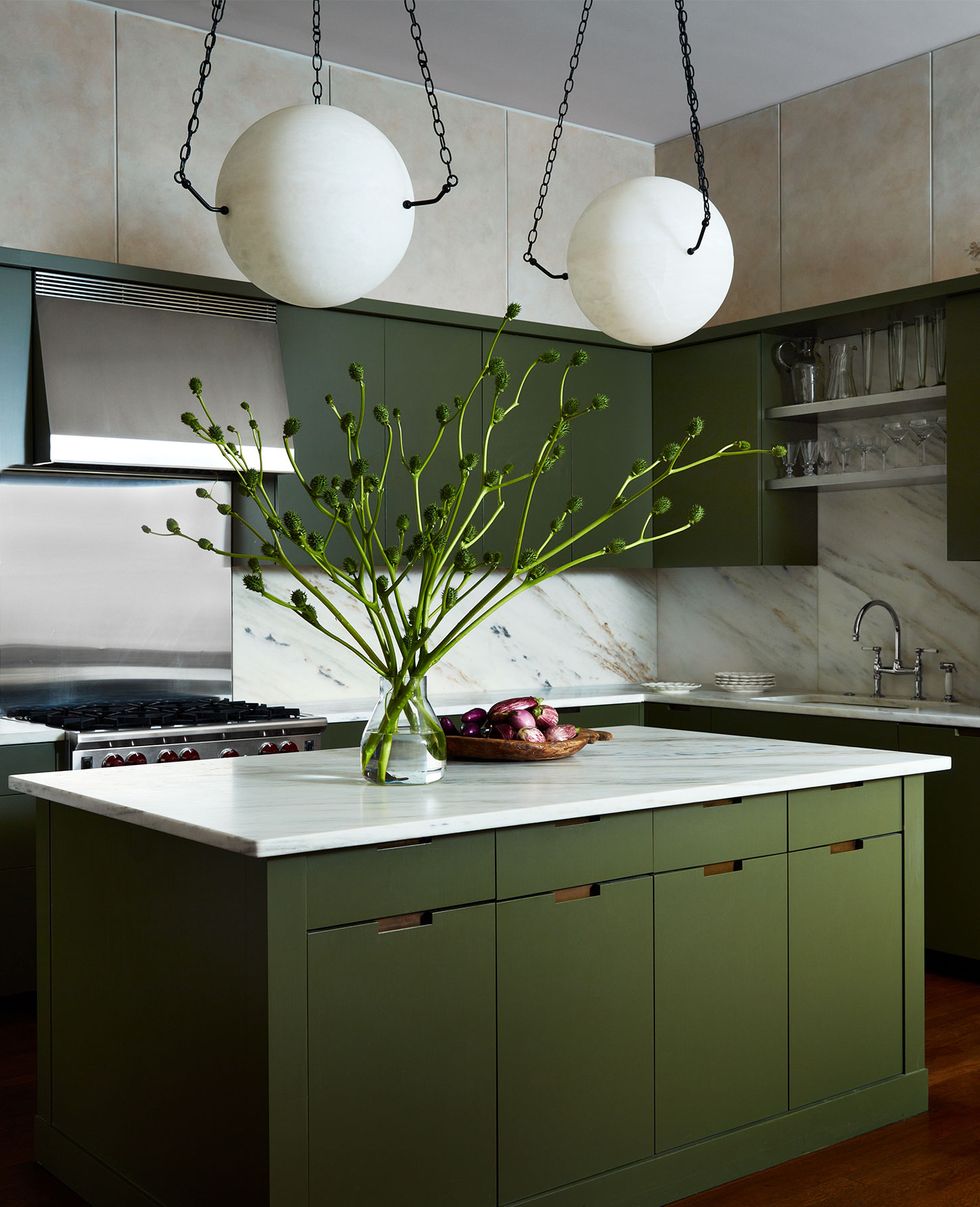 a kitchen has an island with a marble surface and dark olive green cabinets, two alabaster globe pendants suspended by chains, marble backsplashes, stainless oven, sink with open shelving and glasses above