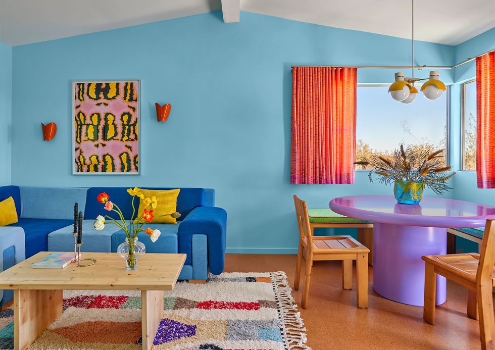 colorful living room with light blue walls and a dark blue sofa and a purple pedestal table and wood chairs and burnt orange curtains on half size window and a colorful rug under the cocktail table