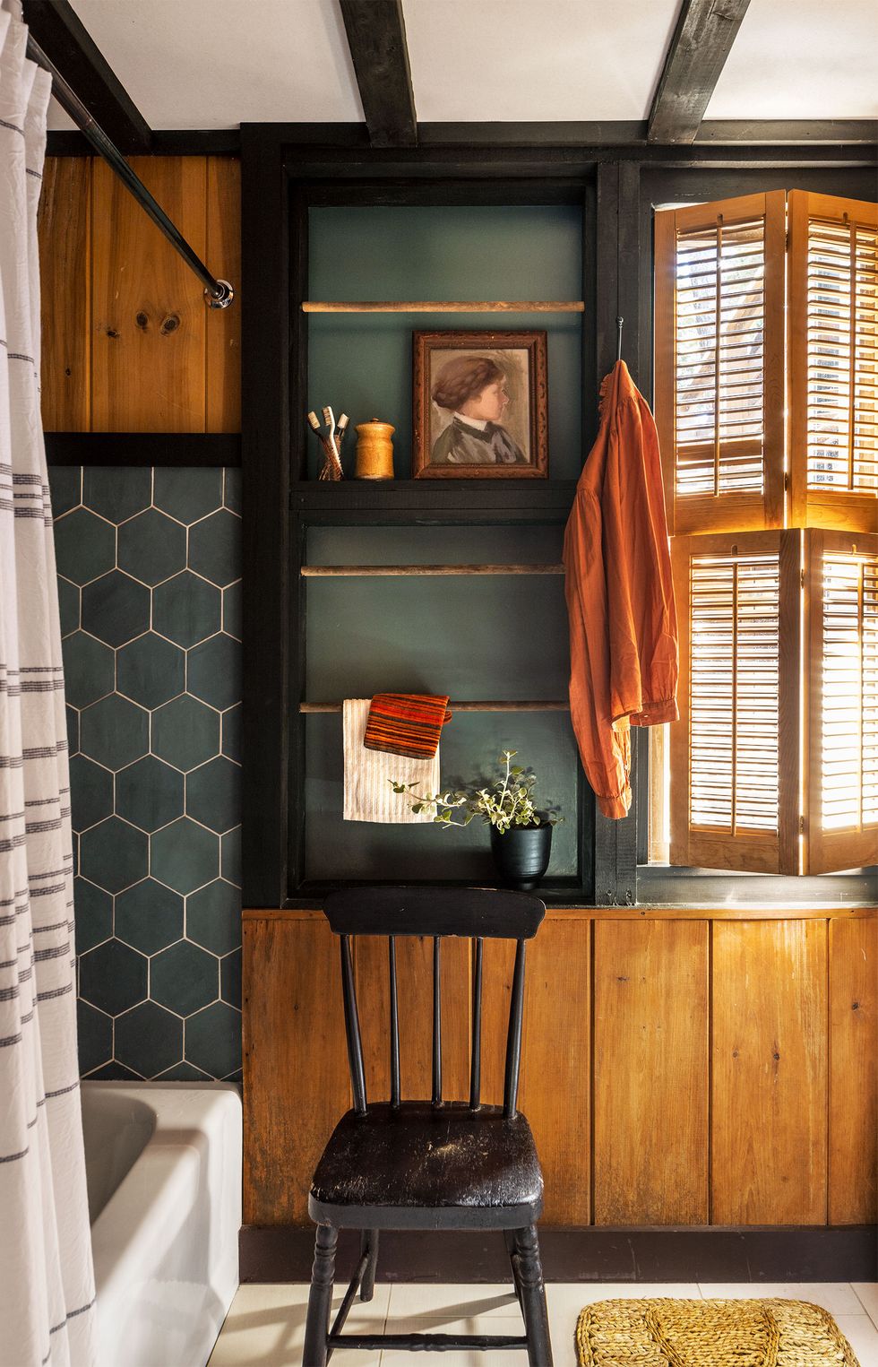 a primary bathroom has dark teal colored hexagonal tiles around the bathtub, a worn black wood chair, wood lower cabinets, a set of inset dowels for towels, a window with wood shutters, and a sisal rug