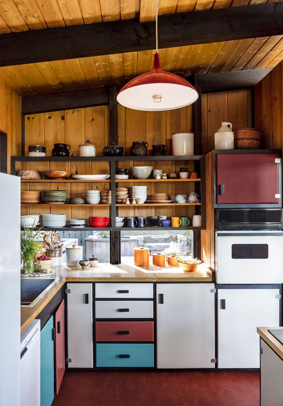 a kitchen has lower cabinets with alternating white, aqua, and deep burgundy colored fronts, open shelving above with plates, cups, and jugs, a red marblelike floor, and a vintage red pendant