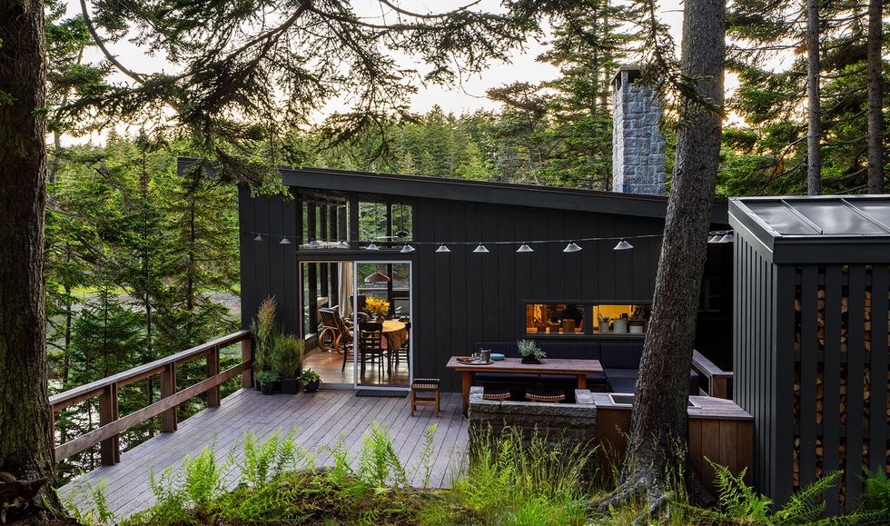 a back deck nestled among evergreen trees above a cove has lamps suspended over a fir dining table surrounded by chairs, a sink and a stone fireplace, a wood railing, and sliding glass doors to the house