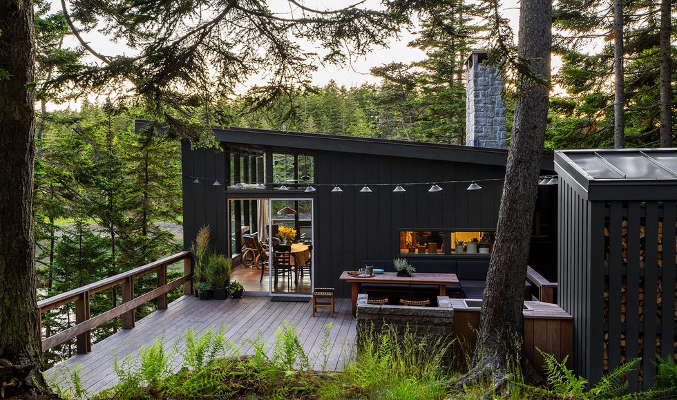 Angie Hranowsky - ELLE DECOR a back deck nestled among evergreen trees above a cove has lamps suspended over a fir dining table surrounded by chairs, a sink and a stone fireplace, a wood railing, and sliding glass doors to the house