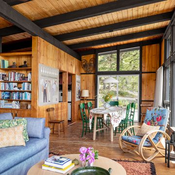 a living and dining area has a blue sofa with accent pillows, a bentwood rocker with a flowered cushion, wood paneled walls and beamed ceiling, a dining table with green painted chairs, a built in book shelf