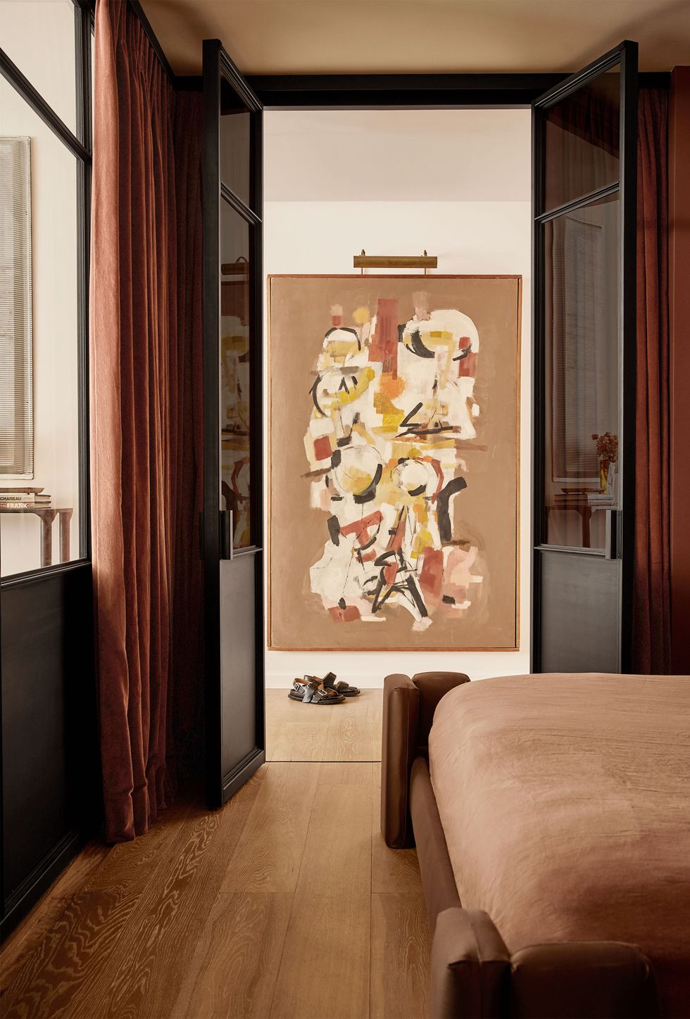 a large modern artwork on a wall faces the to a windowed door to a bedroom that has a wall of frosted glass with terra cotta colored drapes, a bed frame covered in brown fabric and a lighter brown spread