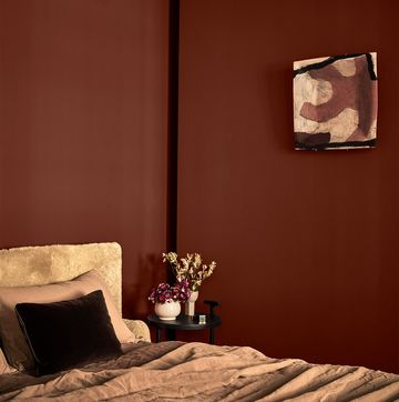 in a brown painted bedroom, a bed's headboard is covered in a light brown shearling, bed covering is in a medium brown linen, a small round night table has a plant and a vase with flowers, a small artwork on wall