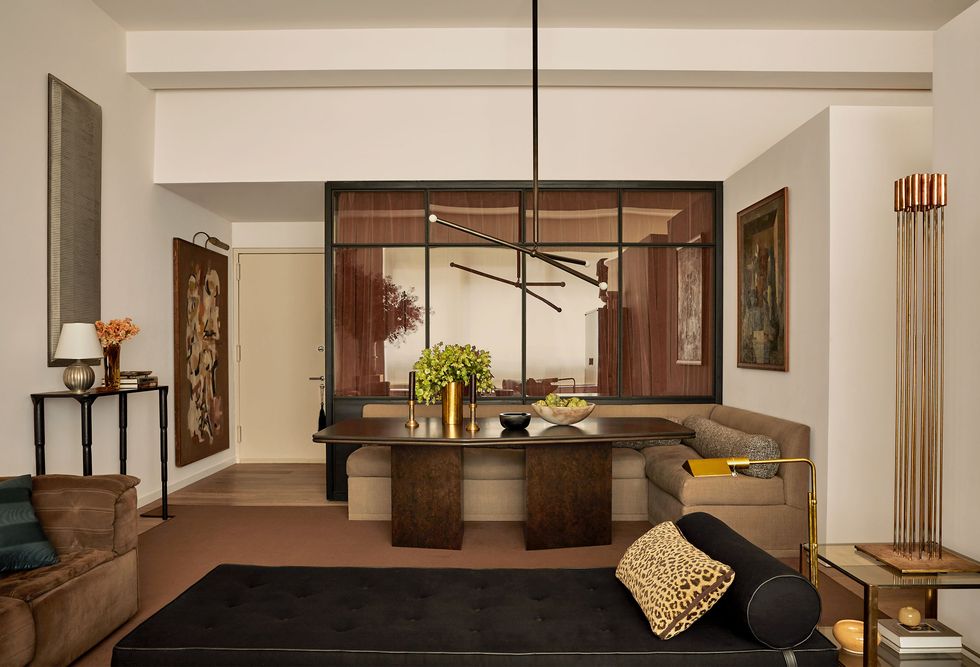a living and dining area has a dark burl wood table, a corner banquette against a frosted glass wall, a three armed pendant above table, a daybed in a dark fabric, an end table, a brown suede sofa opposite, and artworks