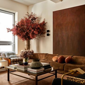 a living room with a long brown suede sofa and a large brown hued artwork above it, cocktail table with books and vases, two chairs with striped cushions and a side table, and a large vase with a huge bouquet of flowers