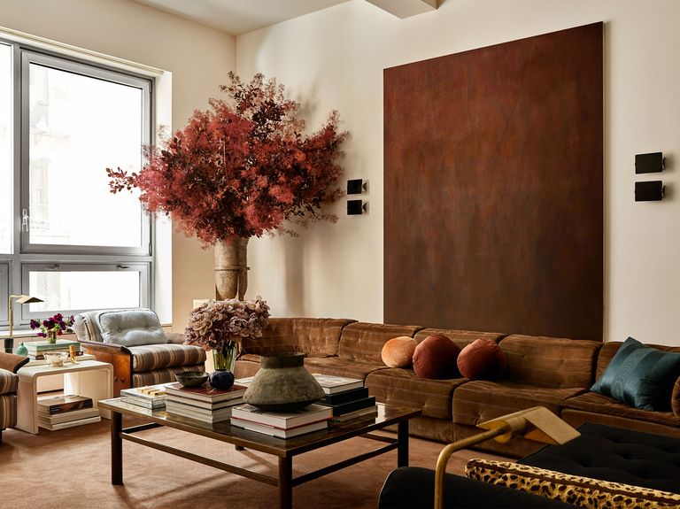 a living room with a long brown suede sofa and a large brown hued artwork above it, cocktail table with books and vases, two chairs with striped cushions and a side table, and a large vase with a huge bouquet of flowers
