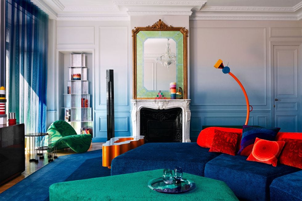 a brightly colored living room by the design brand uchronia, with funky furniture, a white mantel, and a gilded mirror sitting on top of the fireplace