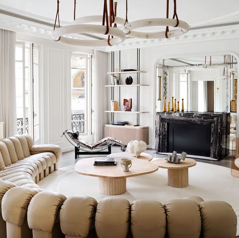 a white room, decorative molding, a trio of circular pendants, semicircle sofa in tufted beige leather, round rug, three cocktail tables, lounge chair, acrylic chair with cylindrical seat, black marble fireplace