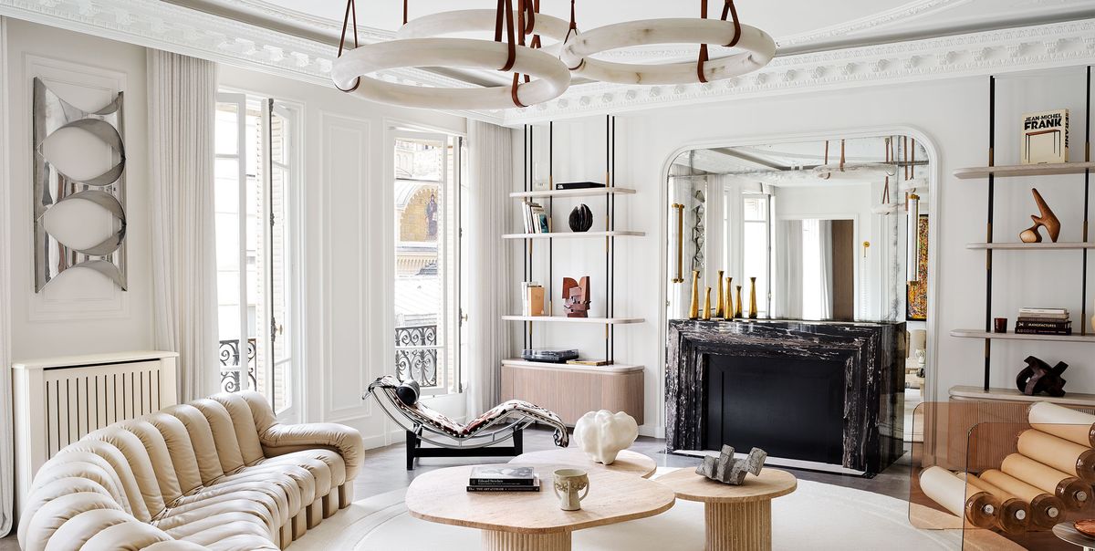 a white room, decorative molding, a trio of circular pendants, semicircle sofa in tufted beige leather, round rug, three cocktail tables, lounge chair, acrylic chair with cylindrical seat, black marble fireplace