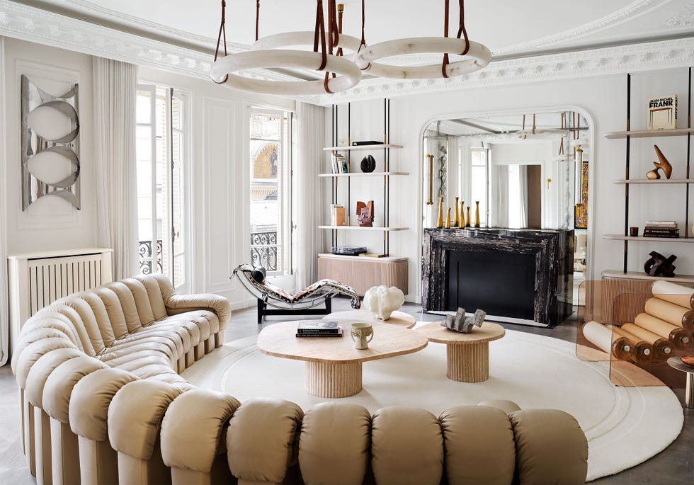 White room, carved moldings, three round pendants, half-round sofa in tufted beige leather, round rug, three cocktail tables, lounge chairs, acrylic chair with stacked cylinders for seating, black marble fireplace