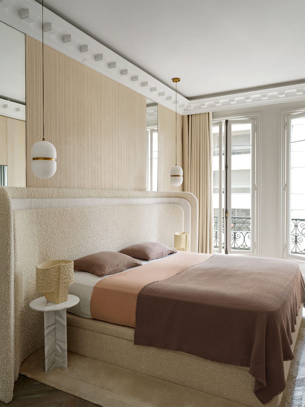 Sarah Dray - ELLE DECOR a bed with upholstered curved headboard and frame with brown and peach bedding, marble nightstands with lamps, grooved wall treatment, suspended pendants, curtained windows to a small terrace