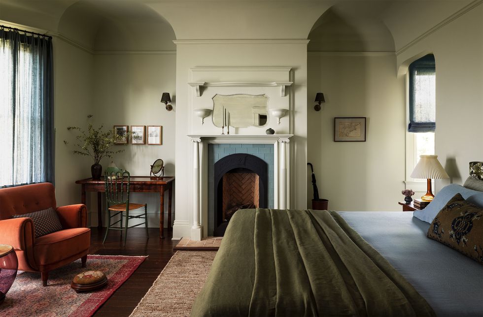 bed has slate blue sheets and an olive green blanket, upholstered armchair, two arched niches flank a fireplace, one niche with writing desk and chair, sconces, curtained windows, two rugs