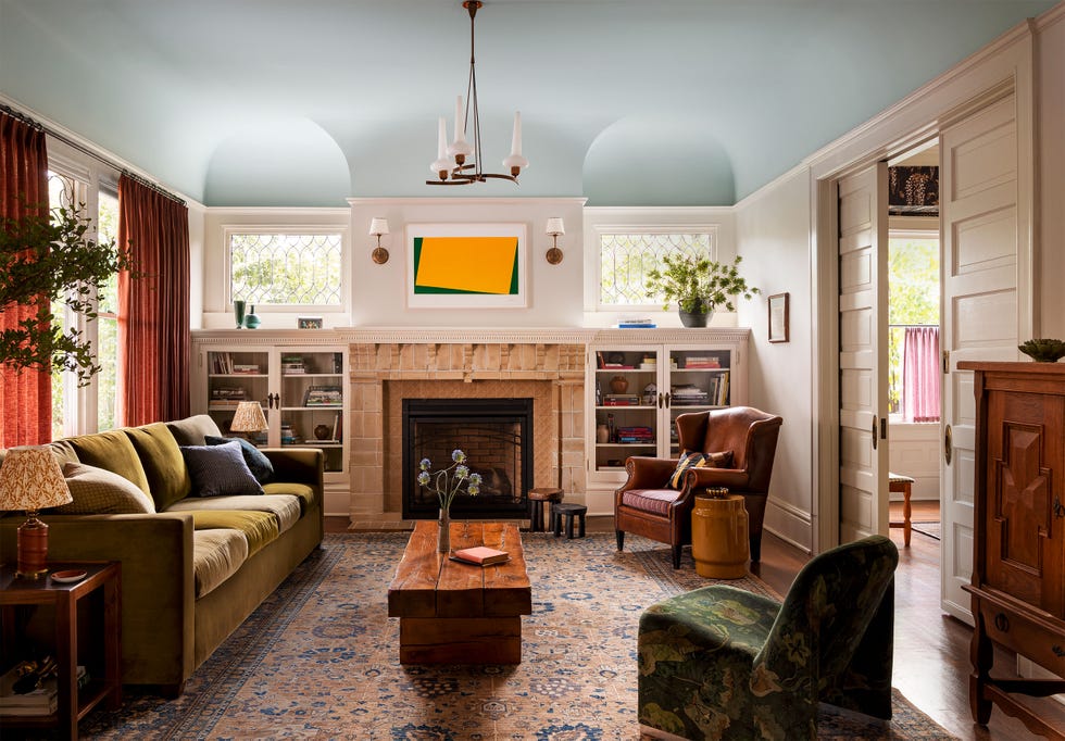 ocher velvet sofa in front of curtained windows in a rust color, bookshelves flank a fireplace with small leaded windows above, leather armchair, slipper chair, cocktail table, turkish rug, vintage chandelier