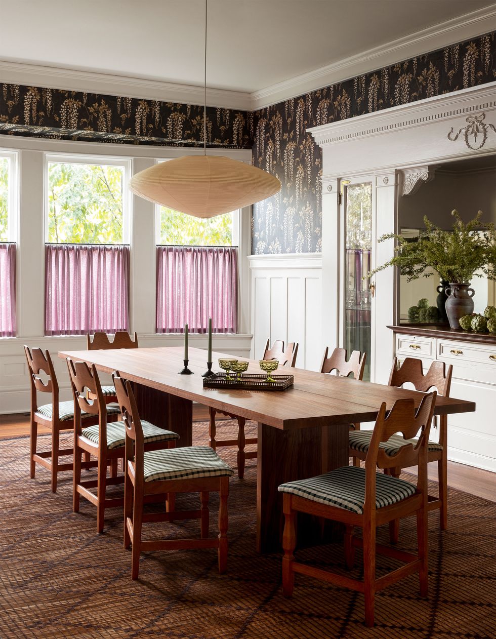 dining room with wood table and eight chairs, paper pendant, walls with white wainscoting and fern print wallpaper above, built in sideboard with mirrored door, windows with half curtains, moroccan rug