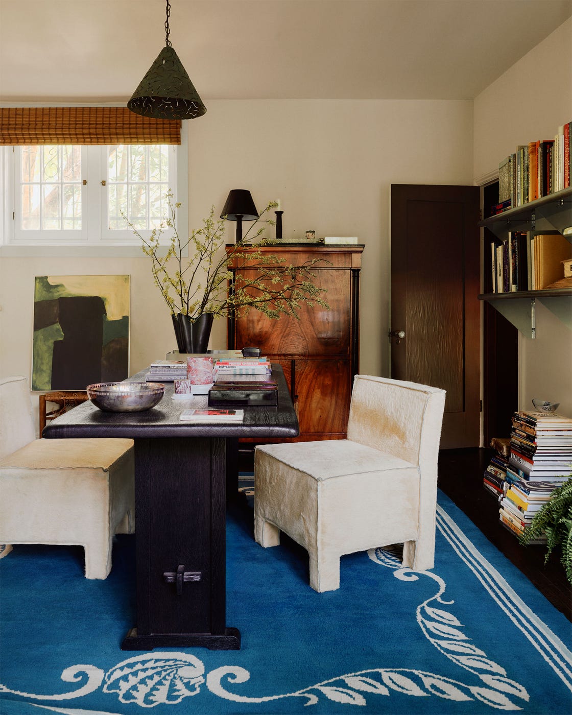 a home office with a dark wood desk and two white square shaped armless chairs, open shelving with books on one wall, closed secretary desk, blue patterned rug