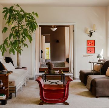 a living room with facing sofas, cocktail table with a sculpture atop between them, small red leather sleigh chair, large potted plant, floor lamps by sofa, end table with shelves, sconce above artworks, bamboo shades
