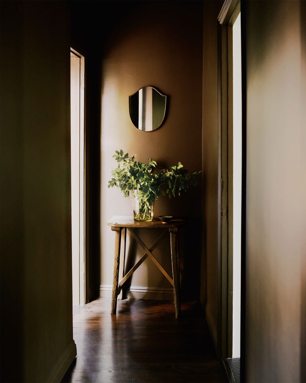 a dark entry all with wood floor, rustic wood stool with a glass vase and greenery, and a shield shaped mirror hanging above