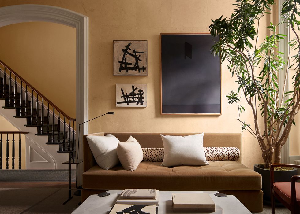 living room with beige sofa against an ocher wall with three framed artworks, large potted plant, cocktail table with light marble top, arched door frame shows a white staircase with black steps and balustrade