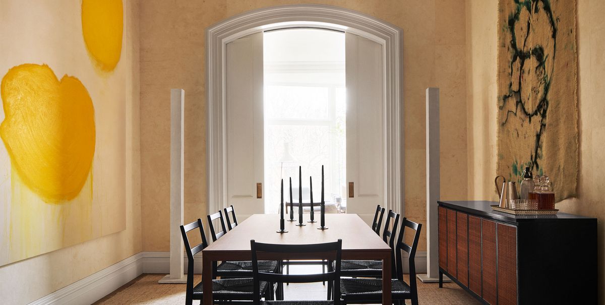 a dining room with ocher colored textured walls, large abstract artworks on facing walls, wood dining table with eight chairs, candelabra, sideboard of dark wood, arched doorway with sliding doors to a terrace