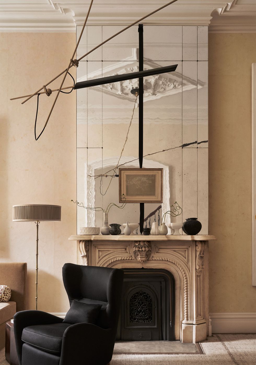 living room of a 1901 building with an fireplace surround and mantel with ceramic vases and a large mirror above it, black wingback chair, floor lamp, black pendant, light ocher walls, neutral carpet