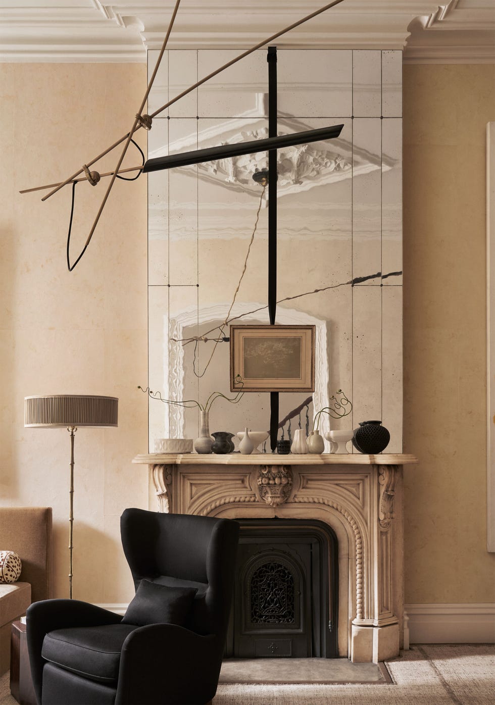 Studio Dorion - ELLE DECOR living room of a 1901 building with an fireplace surround and mantel with ceramic vases and a large mirror above it, black wingback chair, floor lamp, black pendant, light ocher walls, neutral carpet