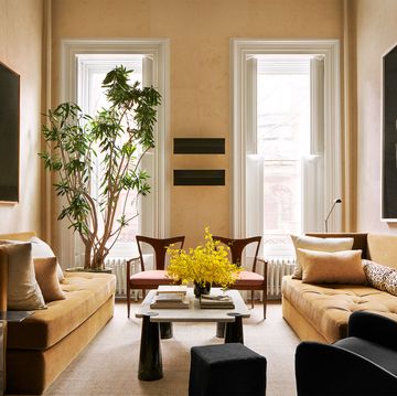 a living room with sofas in a muted mustard velvet, cocktail table with conelike legs, black wingback chair and ottoman, two wooden armchairs, floor lamps, artworks, a plant by tall windows with open shutters