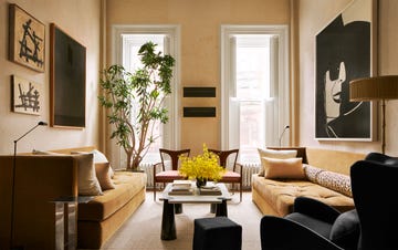 a living room with sofas in a muted mustard velvet, cocktail table with conelike legs, black wingback chair and ottoman, two wooden armchairs, floor lamps, artworks, a plant by tall windows with open shutters