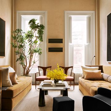 a living room with sofas in a muted mustard velvet, cocktail table with conelike legs, black wingback chair and ottoman, two wooden armchairs, floor lamps, artworks, a plant by tall windows with open shutters
