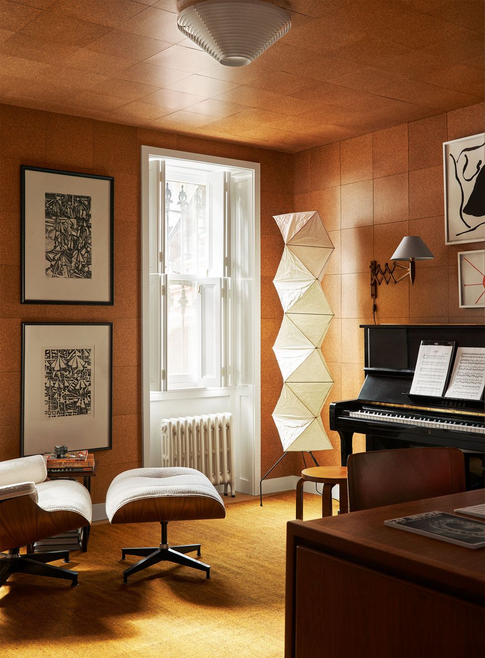 office and music room has a desk and chair, reclining chair with ottoman, an upright piano with accordion arm sconce, artworks above piano and next to recliner, tall paper floor lamp, corks walls and ceiling