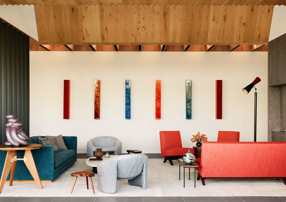 a white room room, slatted wood ceiling, teal sofa and two gray chairs, cocktail table, side table with sculpture, red sofa and two red slipper chairs, floor lamp, wood stool, grouping of six slender rectangular artworks
