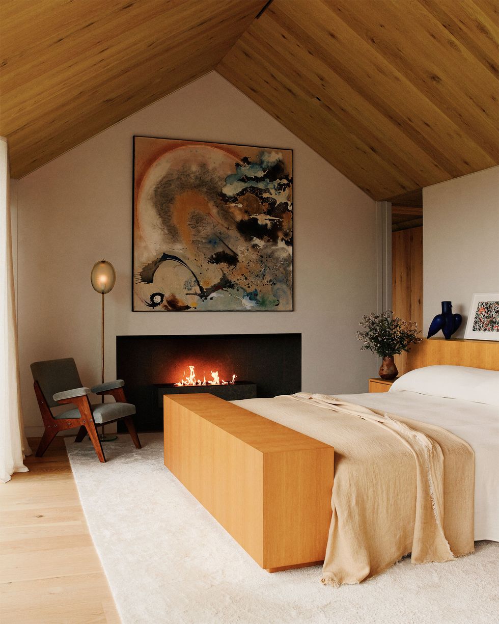 a bed framed in wood with a wide headboard and footboard and attached nightstand with vase, white and blush linen bedding, fireplace with large artwork above, an armchair, floor lamp, light rug, pitched wood ceiling