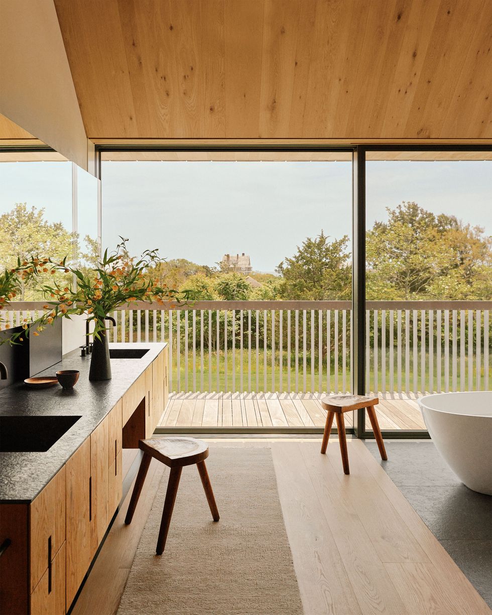 bathroom with floor to ceiling windows looking out to terrace and landscape beyond, double sink vanity with wood cabinets and dark top, runner on wood floor, two wood stools, white soak tub, gray tiled floor under tub